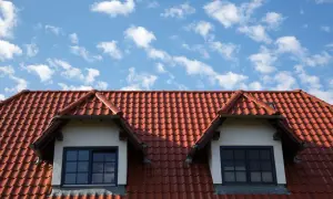 Czerwony dach z dachówki ceramicznej z dwoma lukarnami i niebieskim niebem w chmurach.