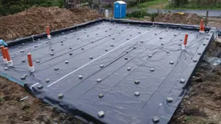 Na budowie widać fundament z folią i liczne dystanse pod zbrojenie. Pomarańczowe rury wystają z ziemi.