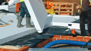 Pracownicy układają płyty styropianu na stropie. Widać cegły, folię i niebieską rurę.