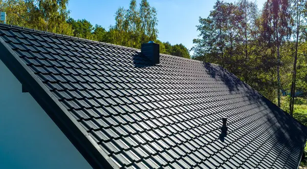 Czarny dach z dachówki ceramicznej z kominem na tle drzew i błękitnego nieba.