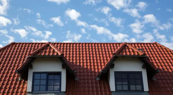 Czerwony dach z dachówki ceramicznej z dwoma lukarnami i niebieskim niebem w chmurach.