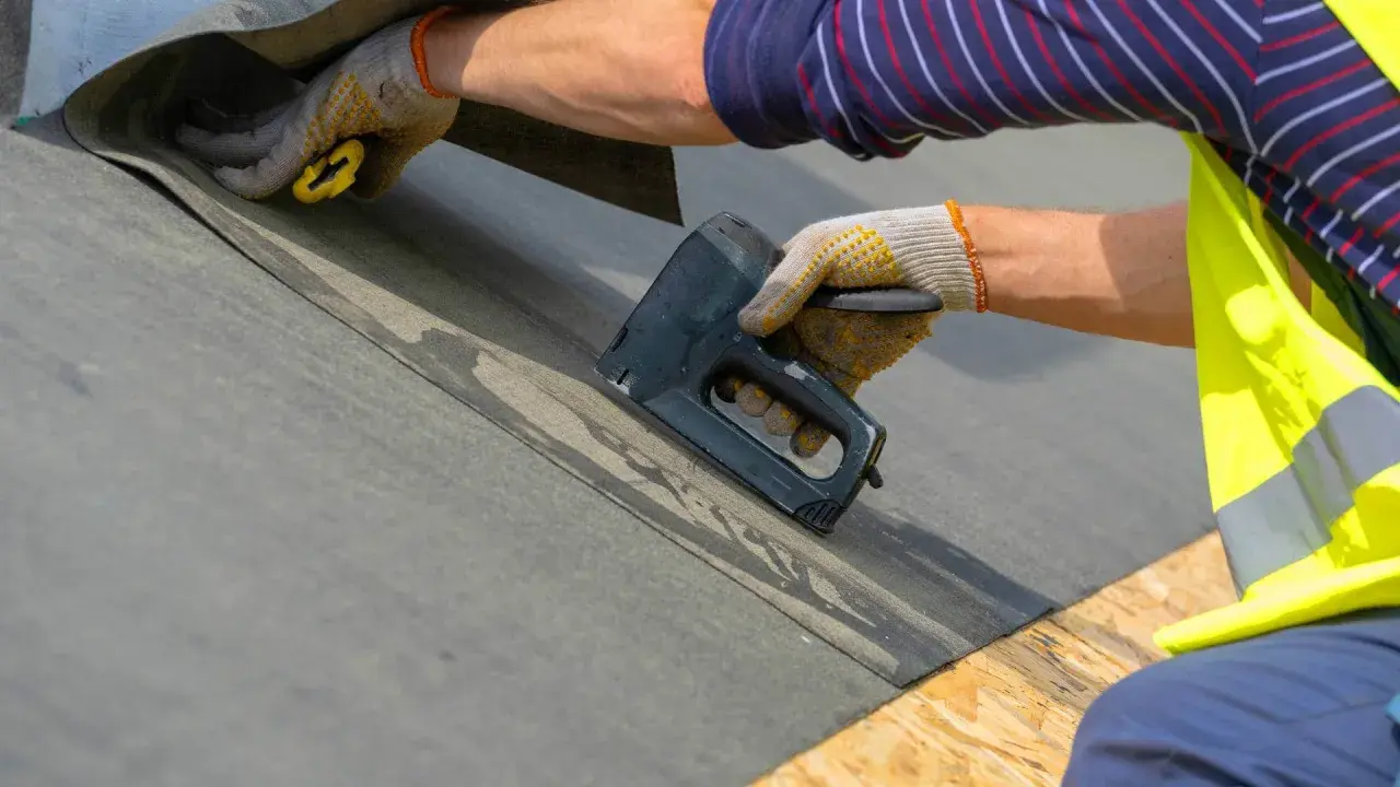 Ręce w rękawicach mocują papę na dachu za pomocą zszywacza.