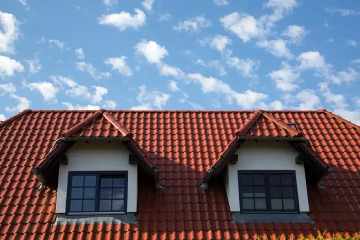 Czerwony dach z dachówki ceramicznej z dwoma lukarnami i niebieskim niebem w chmurach.